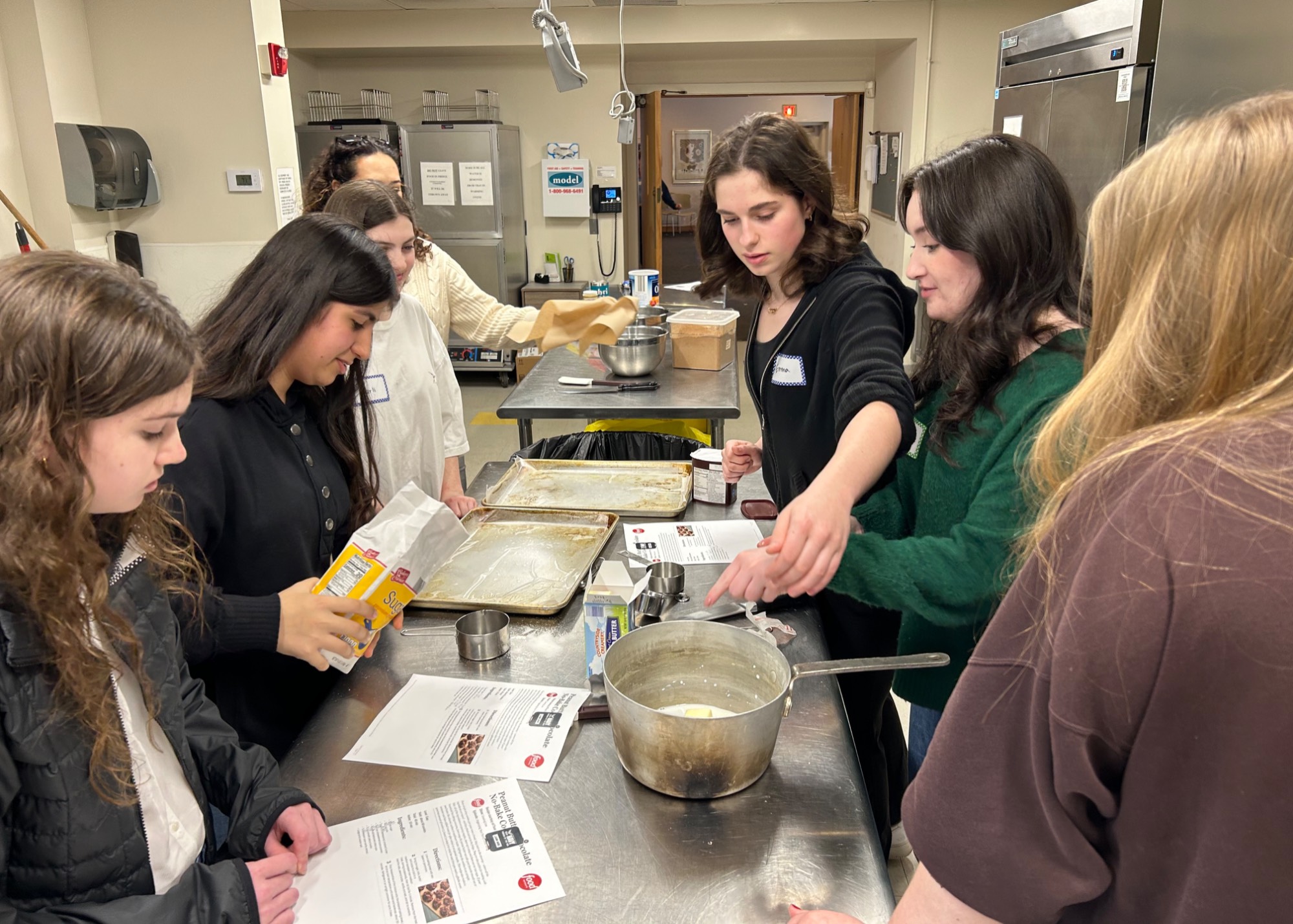 A group of 6 students gathered around an industrial kitchen counter working on a recipe together.